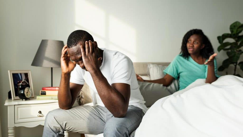 A person sitting on a bed holding their head, with another person gesturing behind them.