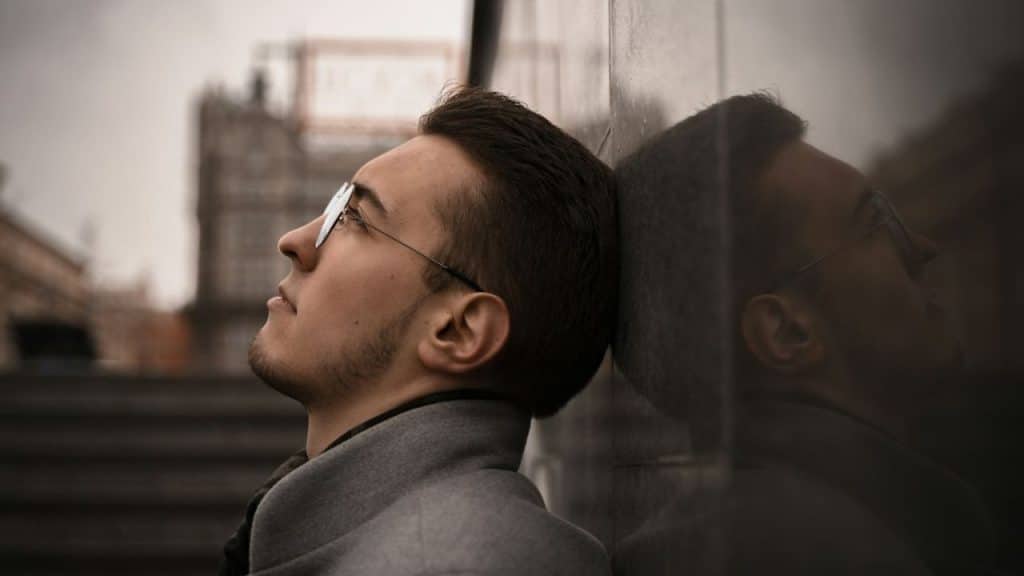 A serious man with glasses and a beard is leaning his head against a stone wall, looking up.