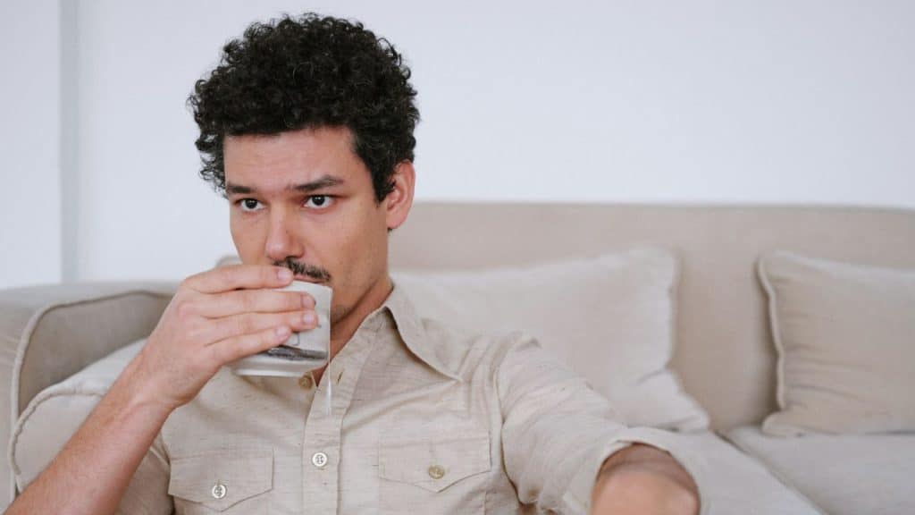 A man with curly hair and a mustache holds a mug to his face while sitting down.