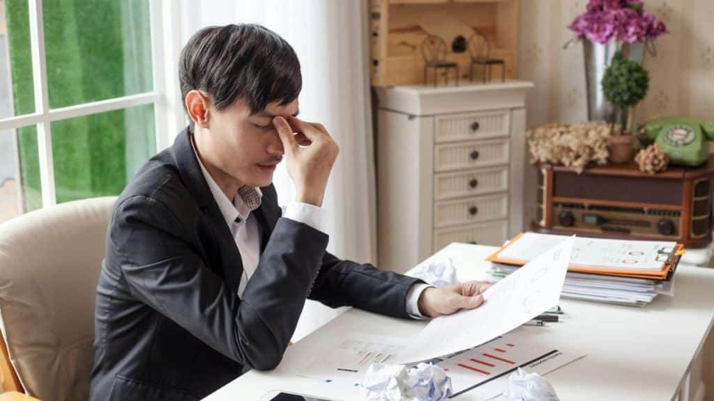 A frustrated man in a suit is at a desk, looking down at a document, with his hand on his face.