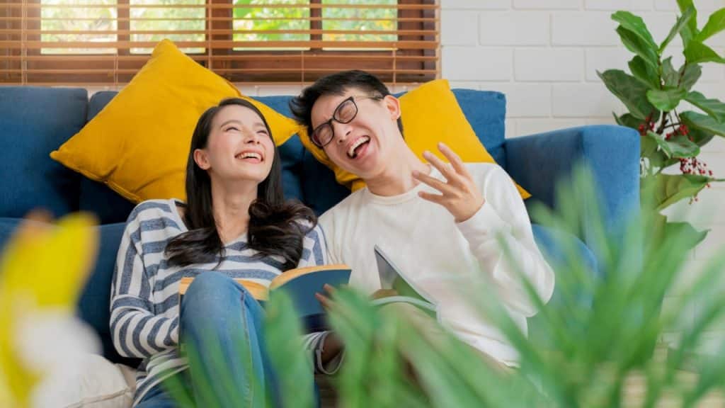 A man and woman laughing while sitting on the floor with books near a blue sofa.