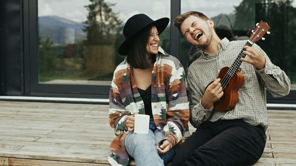 A man plays a small ukulele while a woman in a black hat laughs beside him.