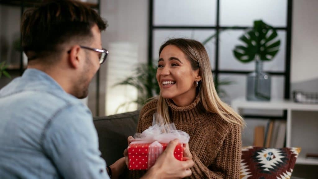 A woman smiles as a man hands her a small, red gift box with a white bow.