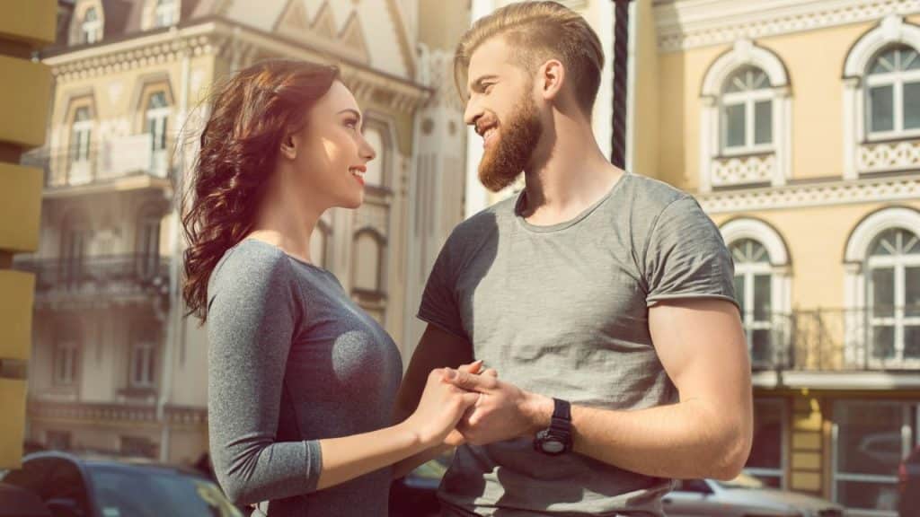 A man with a beard and a woman hold hands while smiling at each other outdoors.
