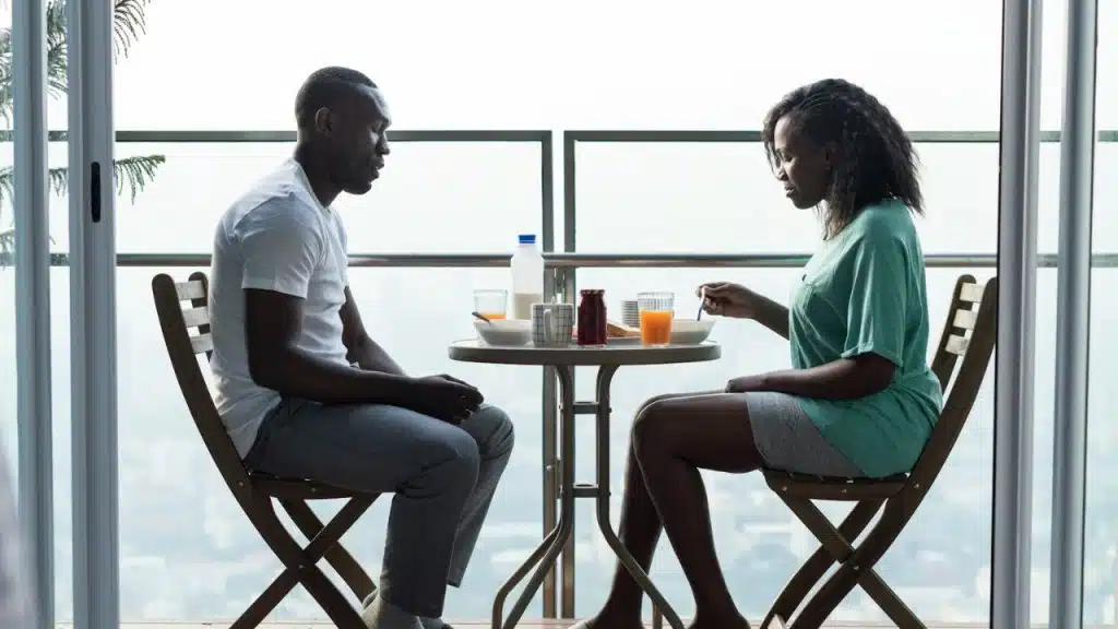 A man and woman sit at a small balcony table with breakfast food and drinks.