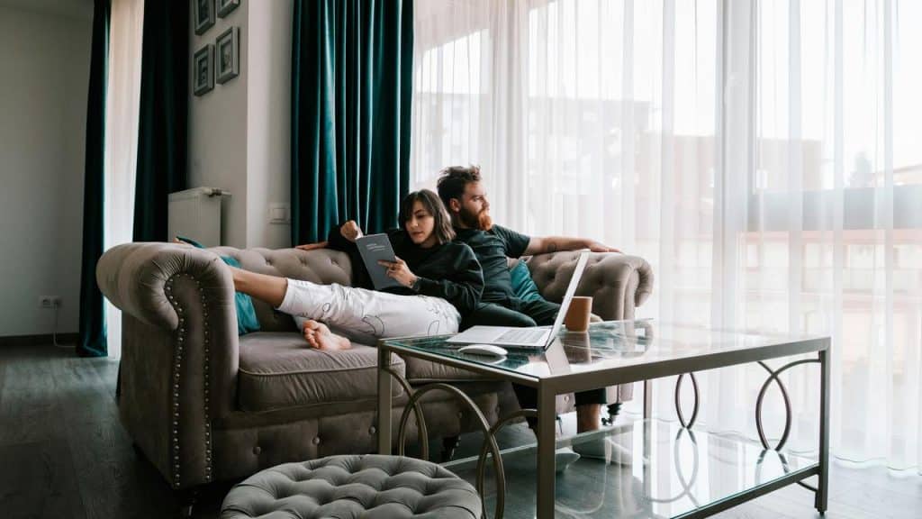 A couple relaxing on a sofa, reading and using a laptop in a bright living room.