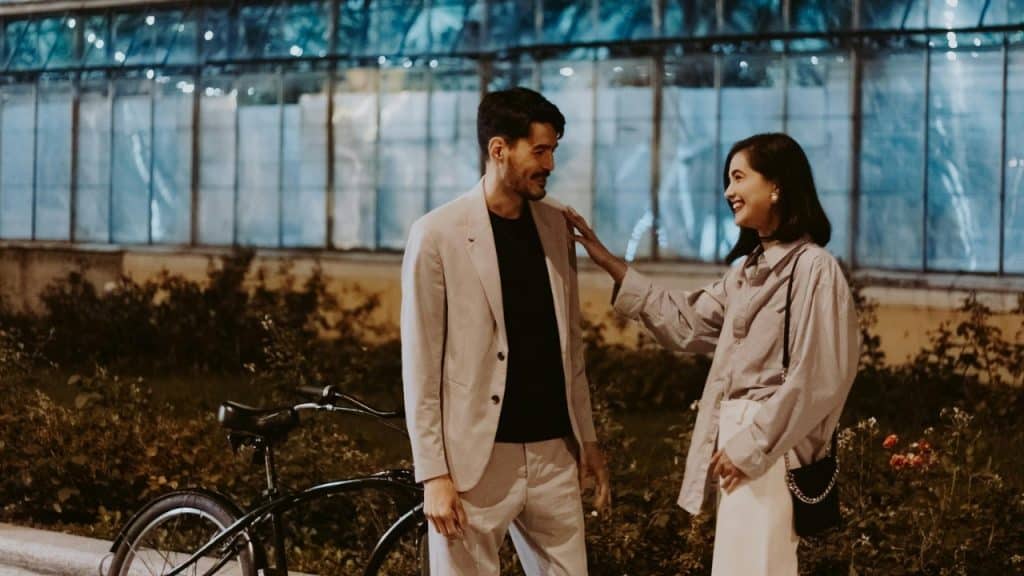 A man and woman stand outdoors at night near a bicycle, smiling and looking at each other.