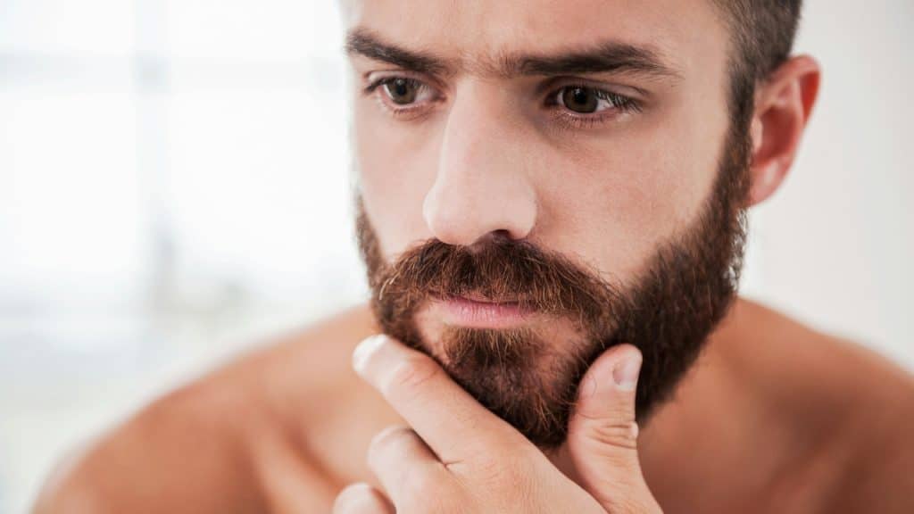 A close-up shot of a bearded man's face with his hand touching his chin.