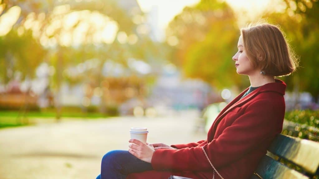 A woman in a red coat sits on a park bench holding a white cup.