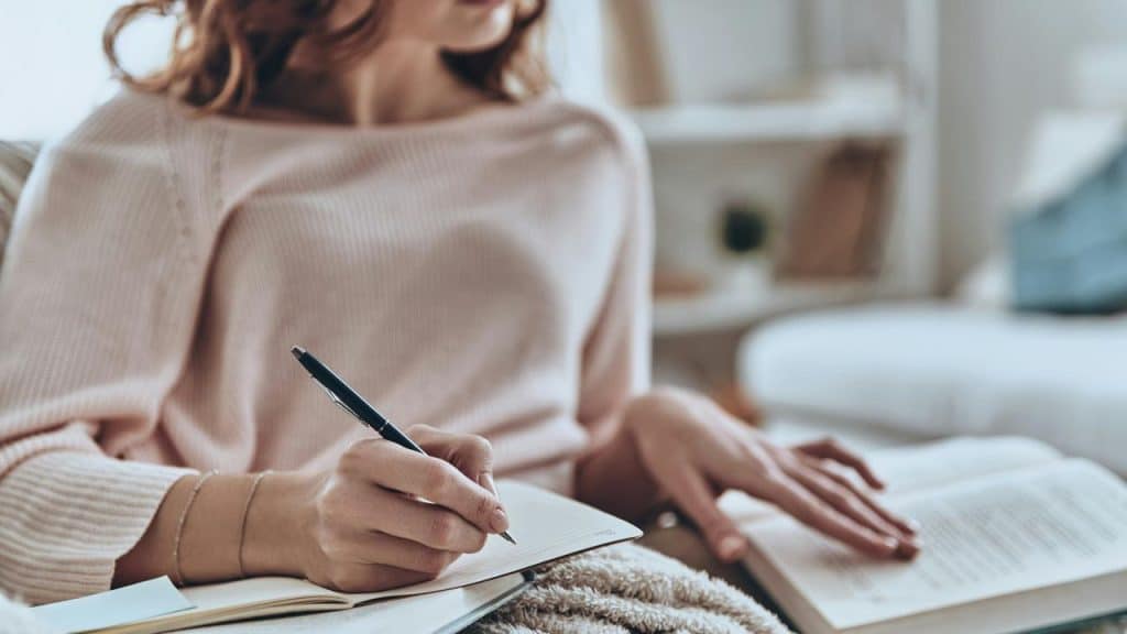 A person writing in a notebook while reading a book in a cozy indoor setting.