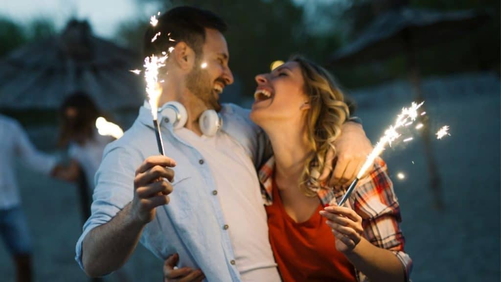 A laughing man and woman embrace on a beach while holding lit sparklers at night.