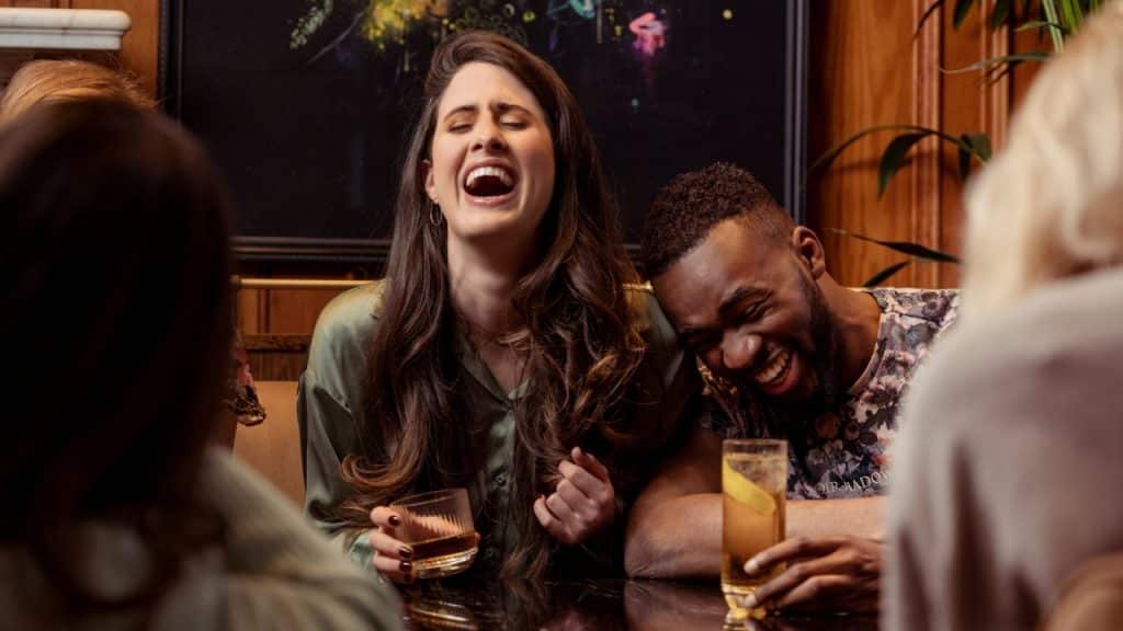A woman in a green shirt laughs while holding a drink next to a laughing man.