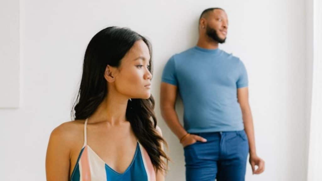 A woman looks away in the foreground while a man stands blurred against a wall.