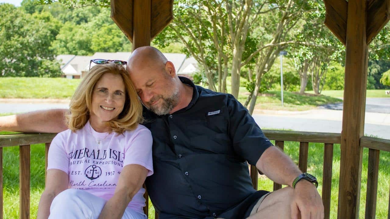 A happy middle-aged couple sitting close together in a wooden gazebo, with the man resting his head on the woman's shoulder.
