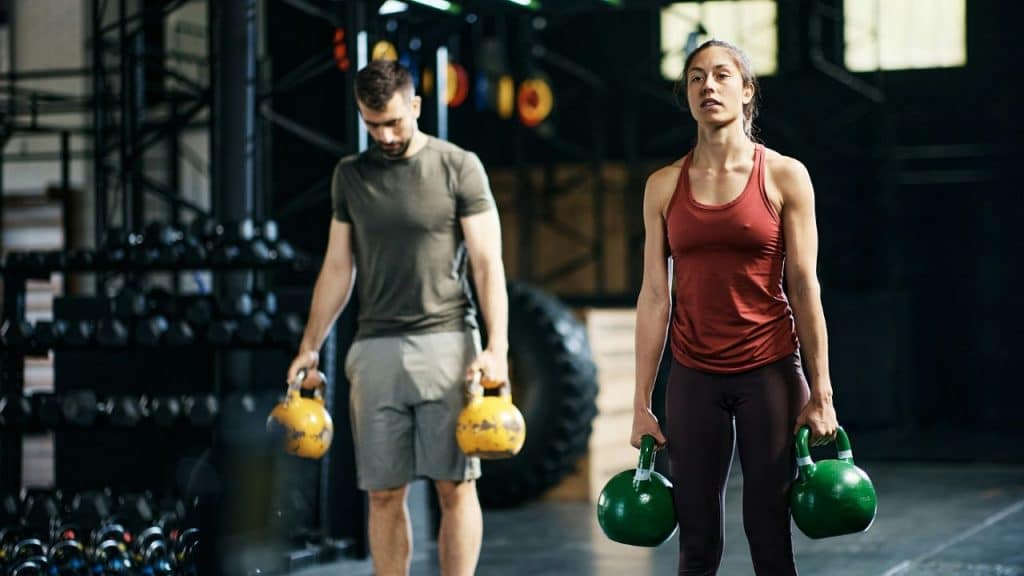 A man and woman stand in a gym holding kettlebells while looking in different directions.