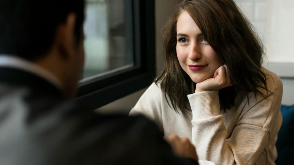 A woman resting her chin on her hand, smiling softly while listening to someone indoors.