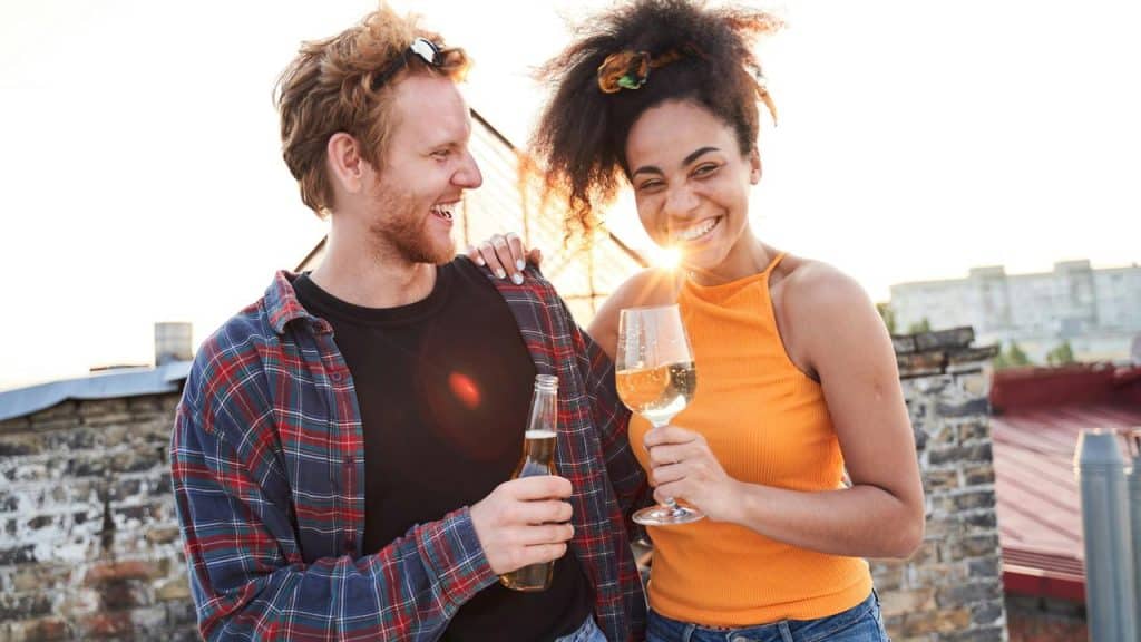 A smiling couple holding drinks together on a rooftop at sunset.