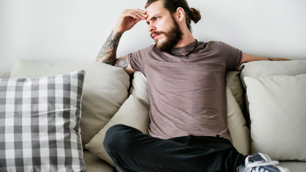 A man sitting on a couch with one hand on his forehead, looking off to the side.