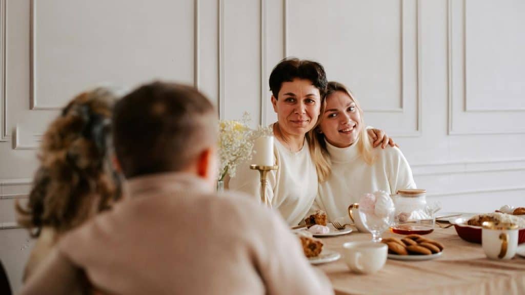 A family gathered around a dining table, with two women smiling and embracing.