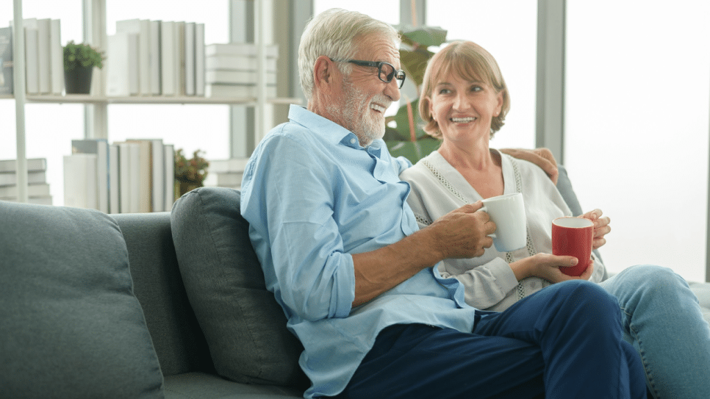 An elderly couple sits on a couch, smiling and holding mugs.