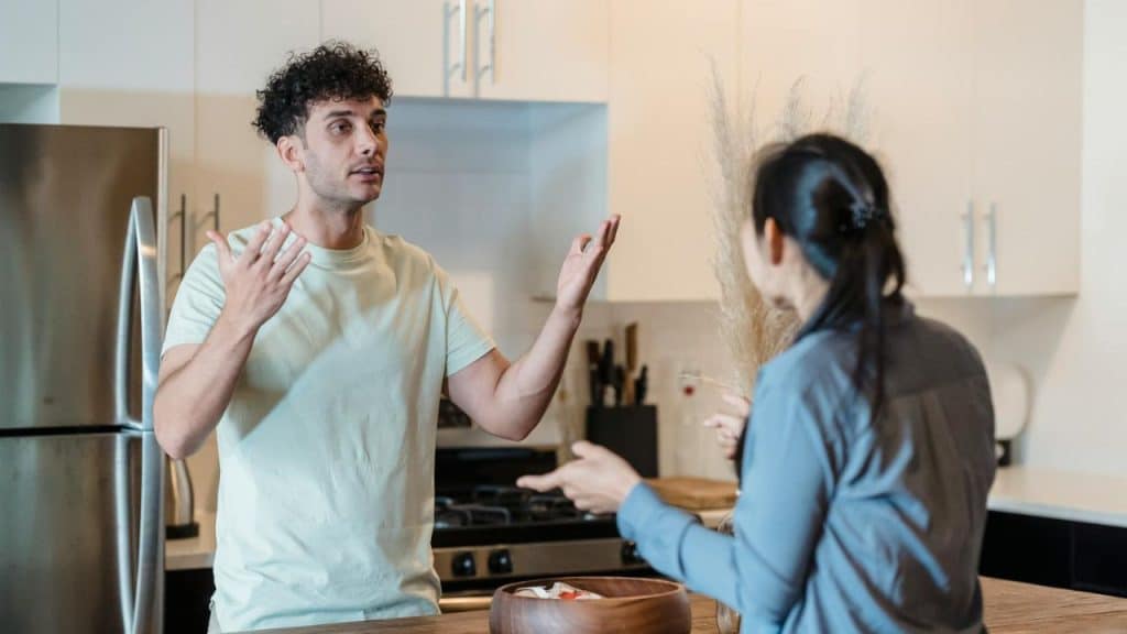 A man with curly hair gestures with both hands while speaking to a woman in a kitchen.