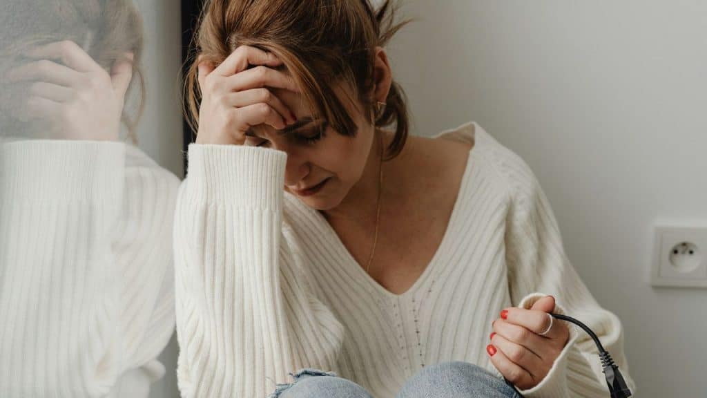 A woman in a white sweater sits with her head in one hand, holding a power cord.