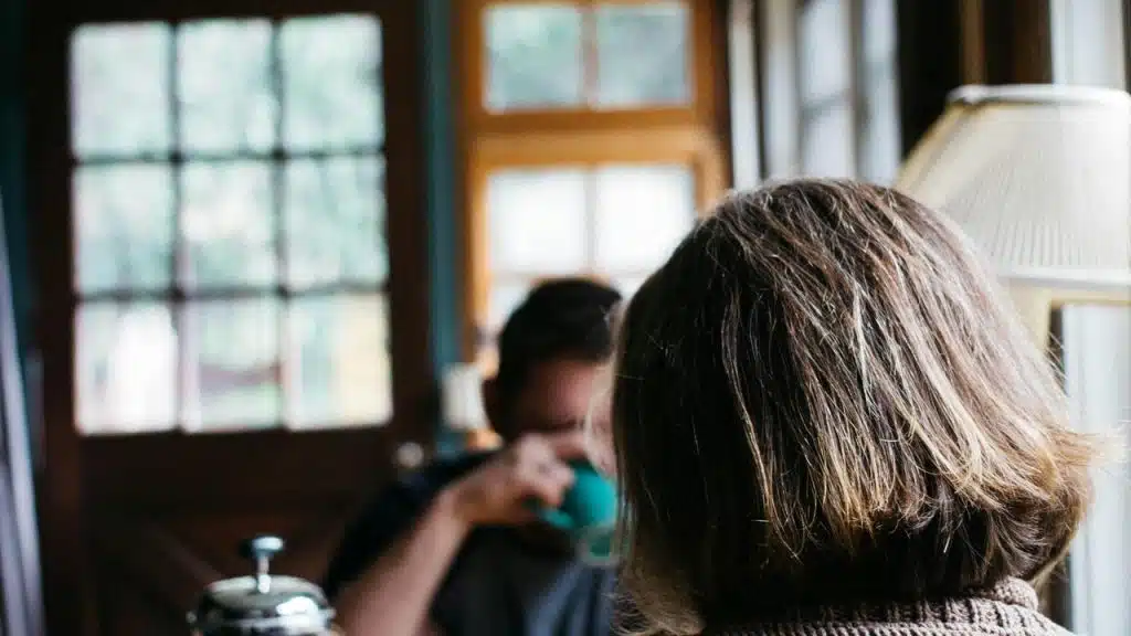 A person sitting indoors with another person blurred in the background near a window.