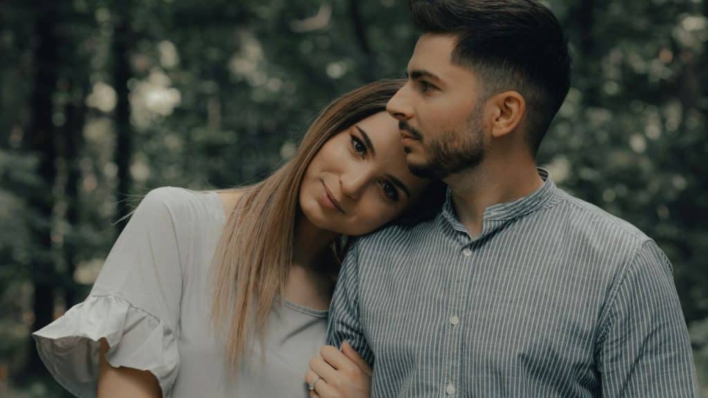 A woman leans her head on the shoulder of a man in a striped shirt outdoors.