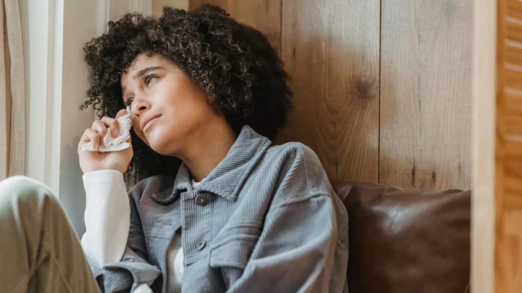 A woman with curly hair sits by a window and wipes tears with a tissue.