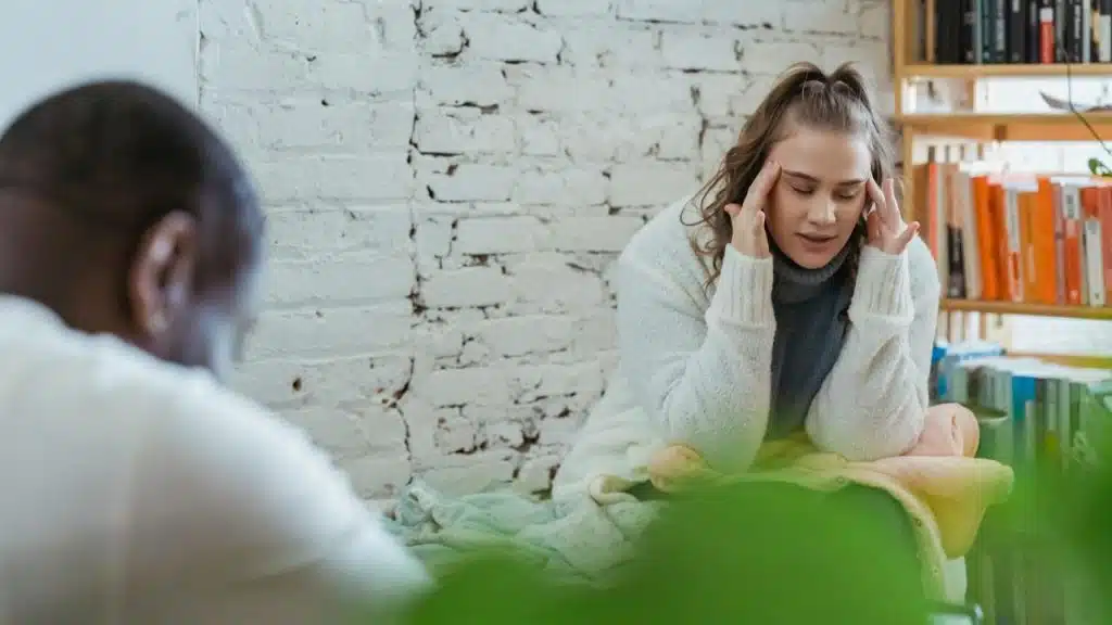 A woman sits on a bed and rubs her temples while facing a man with his back turned.