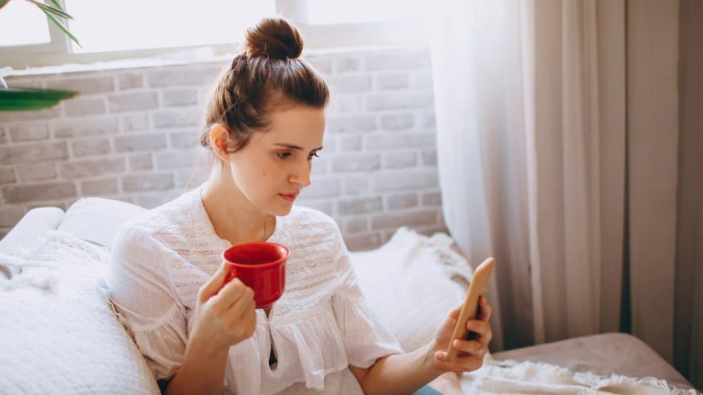 A woman with a hair bun holds a red mug while looking at her smartphone.
