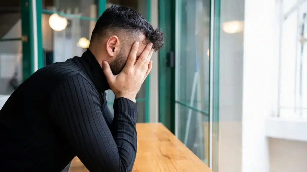 A man in a black turtleneck sits at a wooden table, covering his face with both hands.