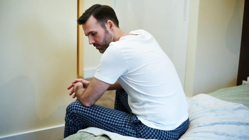 A man in pajamas sits on the edge of a bed, looking down with folded hands.