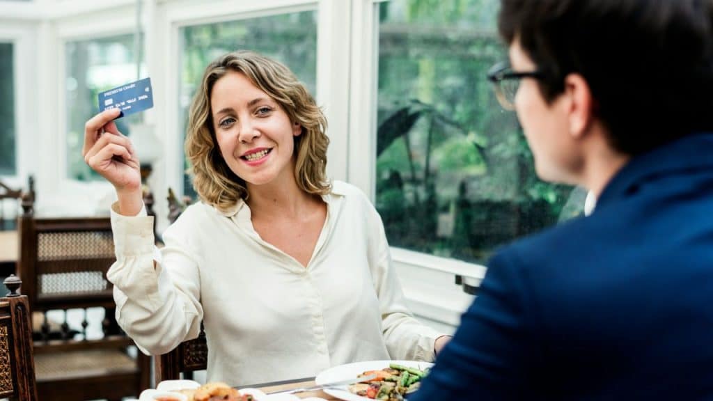 A smiling woman sits at a restaurant table holding up a blue credit card.