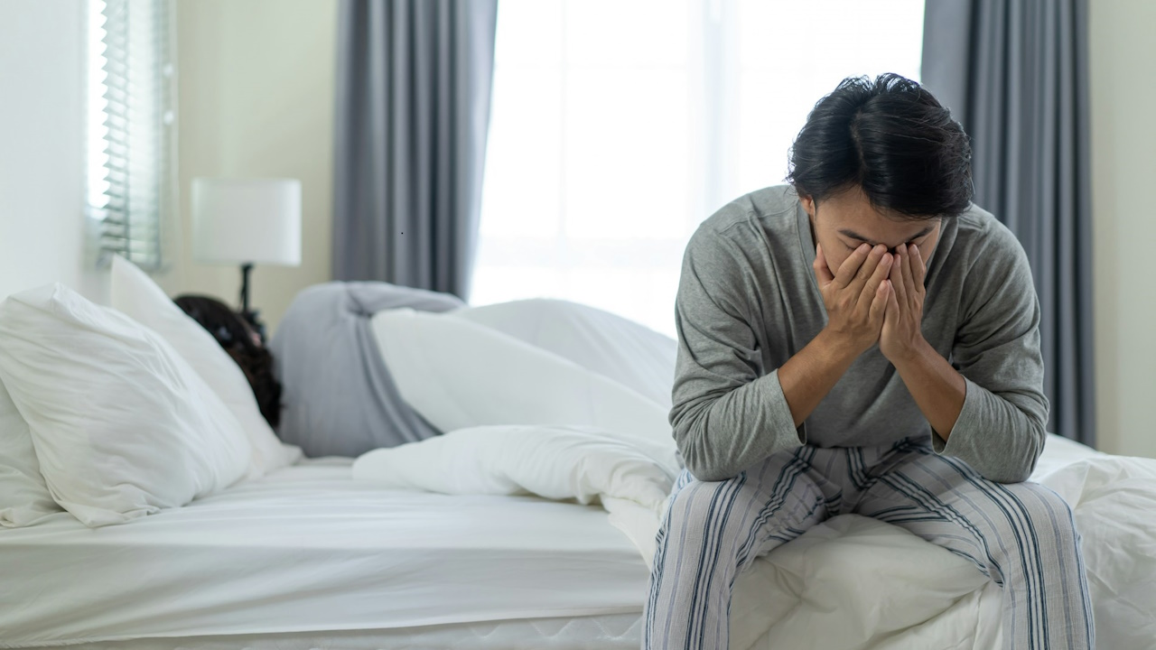 A worried man holding his face in his hands while sitting at the corner of a bed.