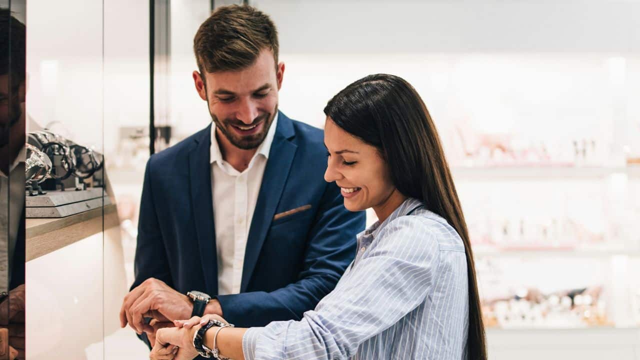 A man and woman smile while looking at wristwatches in a brightly lit jewelry