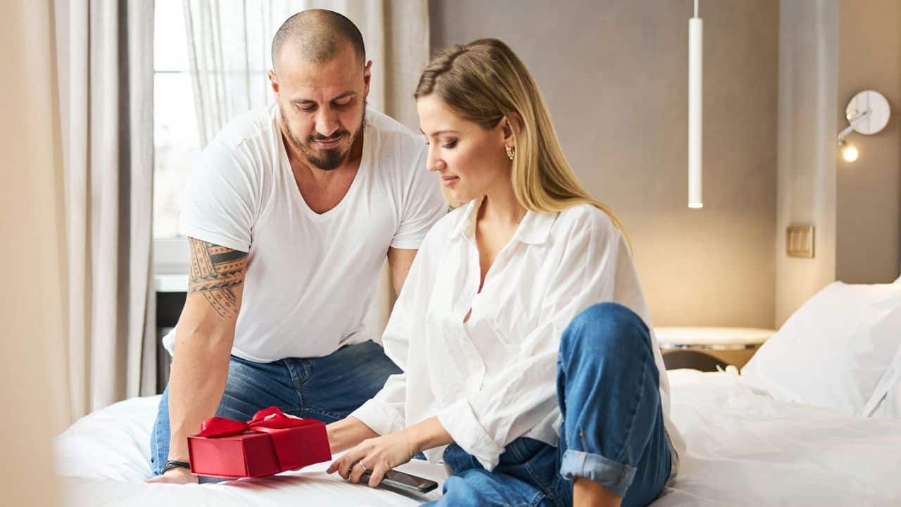 A man and woman sit on a bed together while looking at a red gift box.