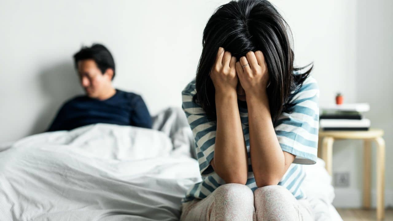 A person sits on a bed with their head in their hands while another person sits behind.