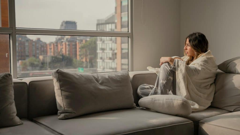 A woman sitting on a couch wrapped in a blanket, looking out a window.