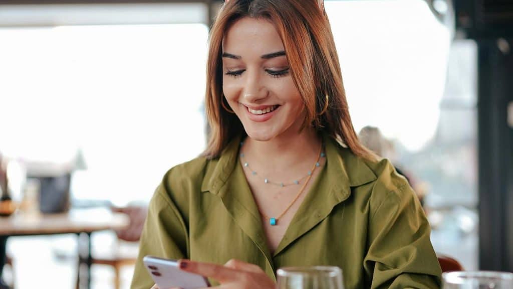A smiling woman in a green shirt looking at her smartphone in a café.