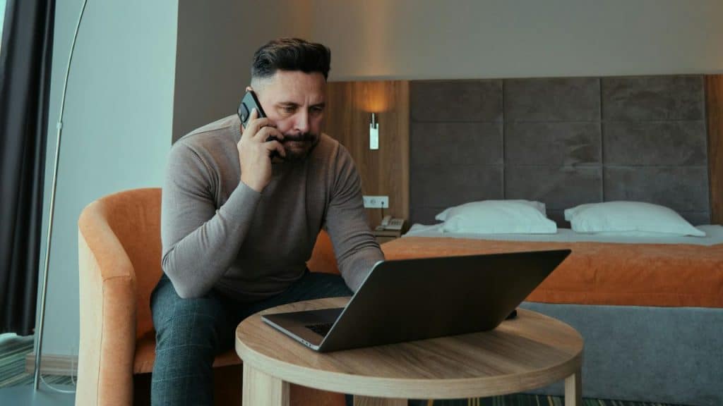 A man talks on a smartphone while sitting at a table with an open laptop.
