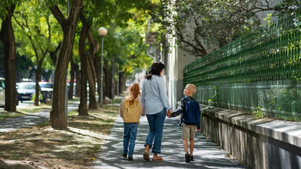 A woman walking hand in hand with two children along a tree-lined sidewalk.