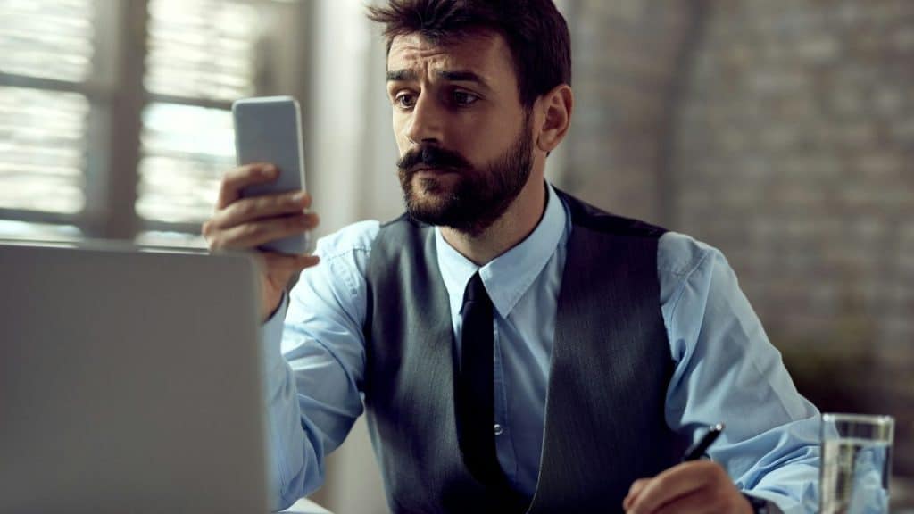A man with a beard and vest looks at a smartphone while holding a pen.