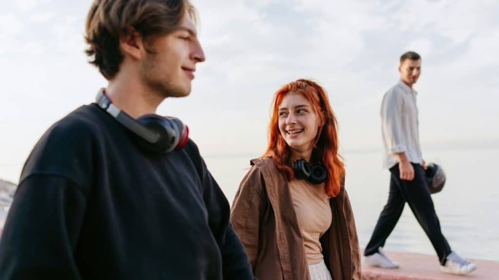 A woman with red hair smiles at a man while another man walks in the background.