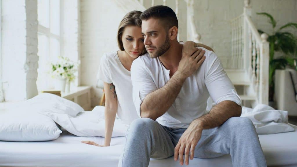 A woman leans her head on a man’s shoulder while they sit together on a bed.
