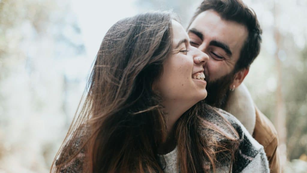 A bearded man kissing a smiling woman’s cheek while holding her from behind.