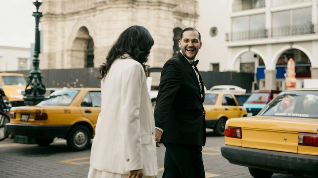 A man and woman in formal attire hold hands while walking past yellow city taxis.