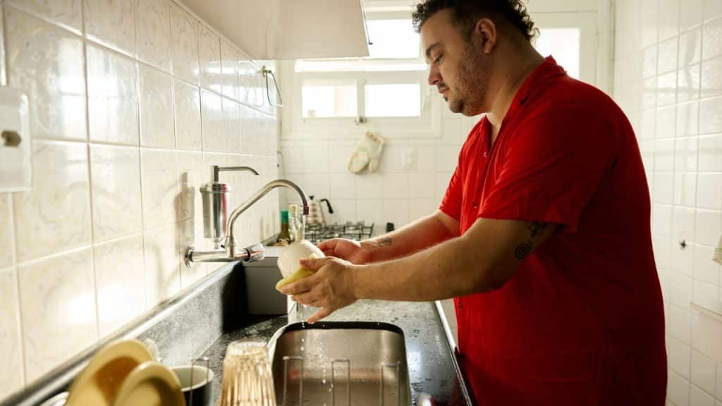 A man in a red shirt washes a white mug at a kitchen sink.