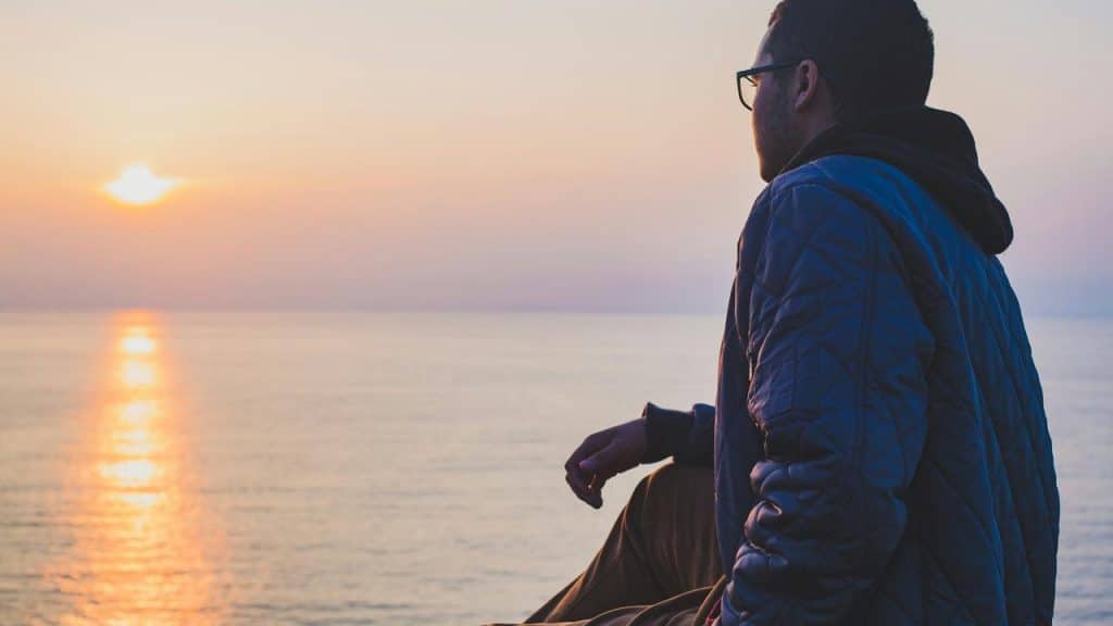 A man sits by the water watching the sun set over the horizon.