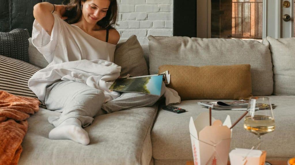 A woman relaxing on a couch with a blanket, a book, and takeout nearby.